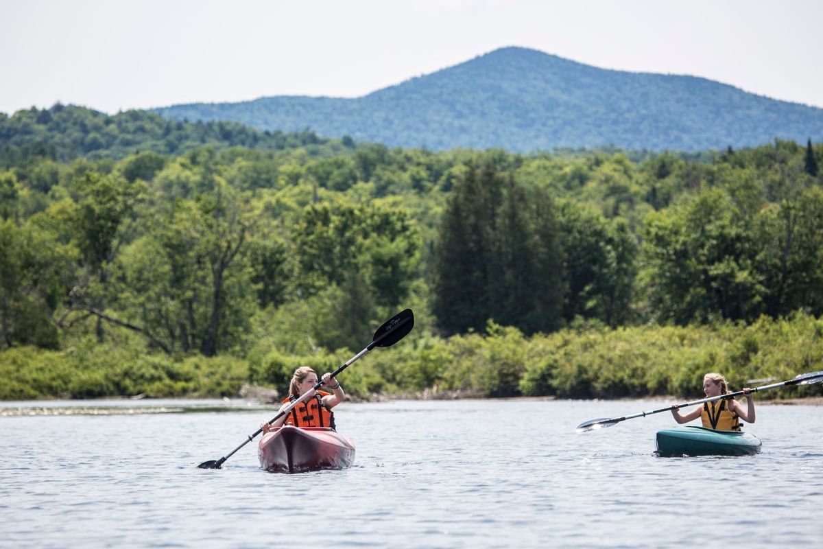 Two People Kayaking On Calm Lake With Life Jackets, Surrounded By Green Trees And Mountain Under Clear Sky